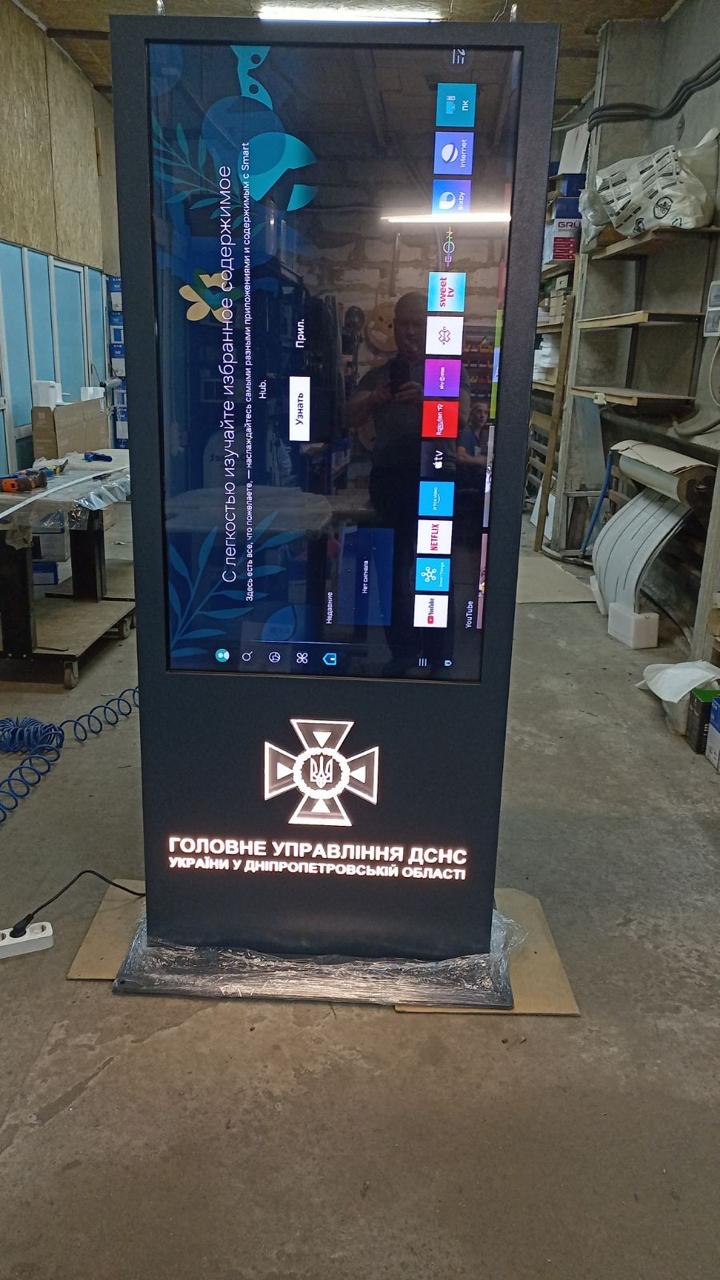 QS55 floor-standing touch kiosk powered on, showing the 55-inch 4K display and illuminated DSNS Dnipropetrovsk regional emblem